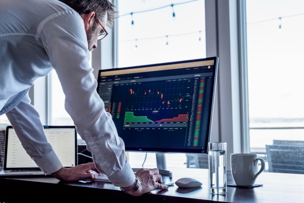 Professional at a wood desk in a bright office, focused on financial charts and data on a large monitor, windows in the background