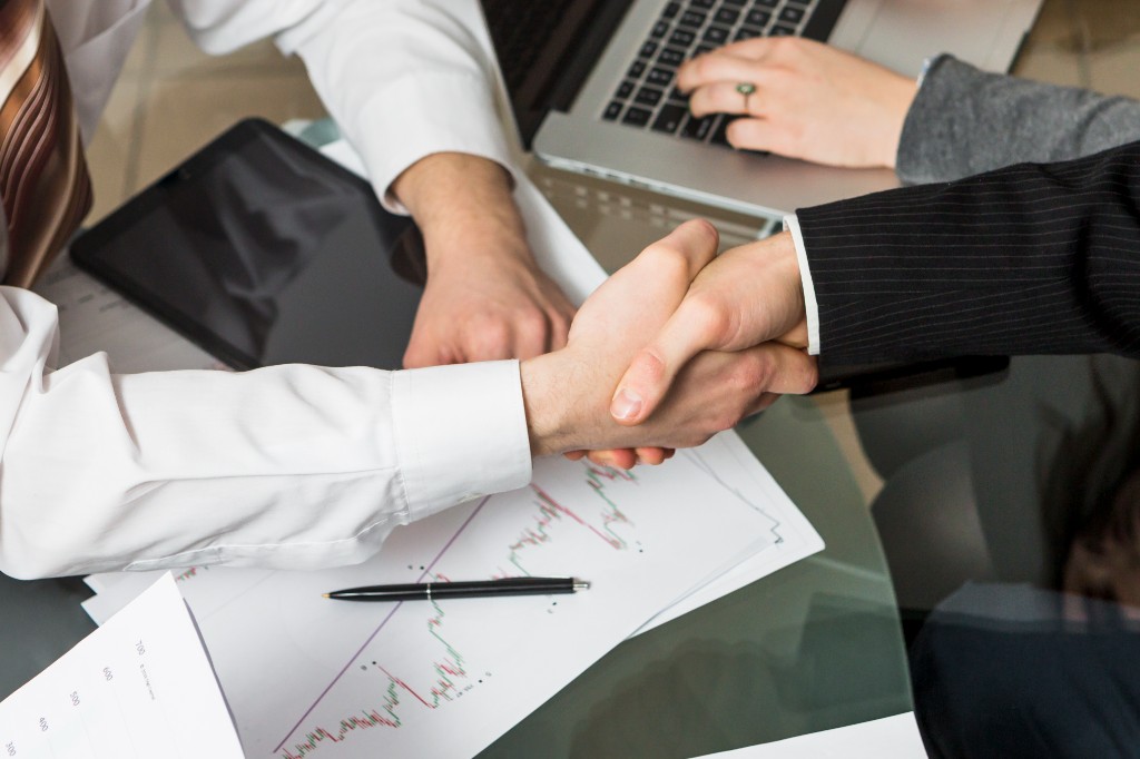 Two businesspeople shaking hands over financial documents, tablet, and laptop on a glass desk