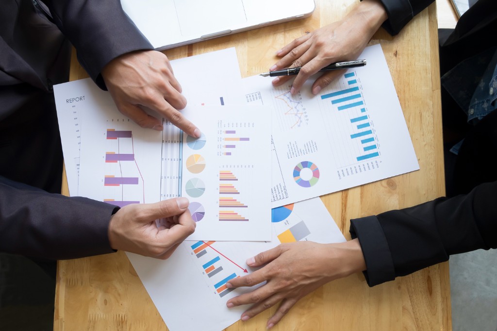 Overhead view of two professionals comparing printed reports with charts on a light wood table